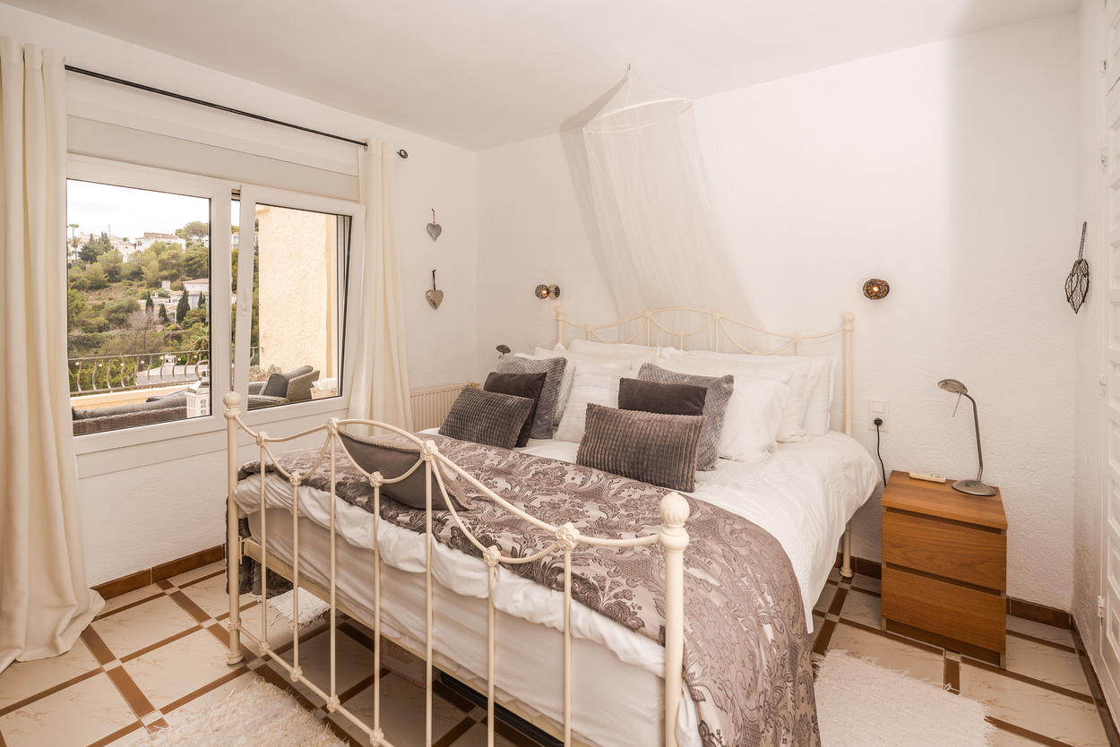 Chambre avec lit en métal blanc, carrelage à motifs et fenêtre double vitrage. La pièce bénéficie d'une lumière naturelle et d'une vue sur les collines.