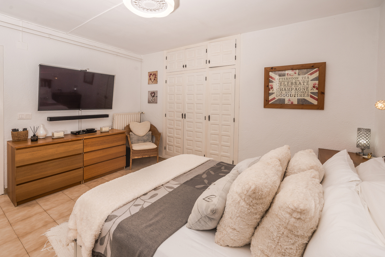 Chambre avec placards intégrés blancs, grand téléviseur mural au-dessus d'une commode en bois et sol en carrelage. Équipée d'un radiateur et d'un fauteuil en osier.