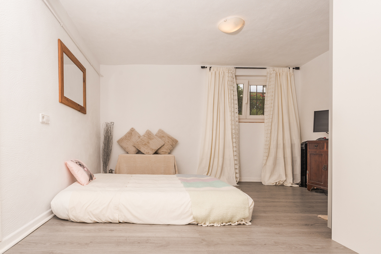 Chambre avec sol stratifié effet bois gris, murs blancs et lit bas. Comprend un meuble en bois sombre, un miroir à cadre en bois et une fenêtre avec rideaux neutres.