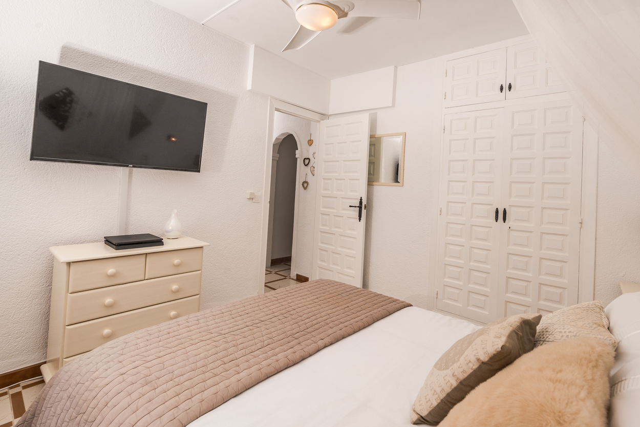 Chambre lumineuse avec placards intégrés à panneaux blancs, télévision murale et ventilateur de plafond. Comprend une commode assortie et un sol en carrelage.