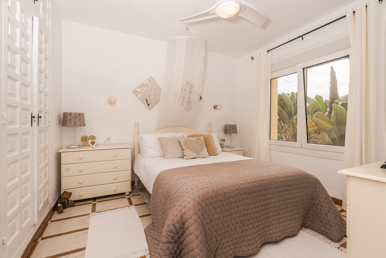 Chambre lumineuse avec placards intégrés, ventilateur de plafond et fenêtres à double vitrage donnant sur le jardin, complétée par un sol en carrelage traditionnel.