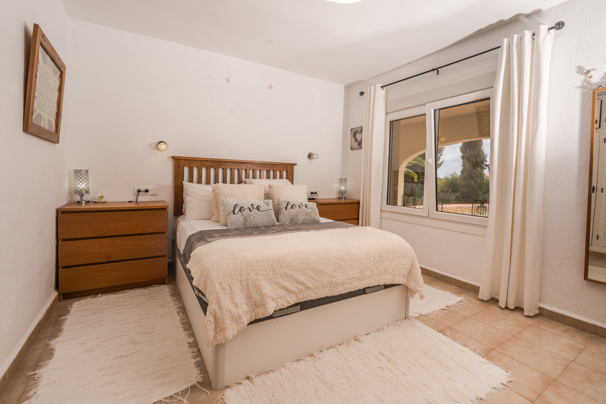 Chambre lumineuse avec tête de lit en bois, fenêtres à double vitrage et sol en carrelage. Comprend des commodes en bois assorties et une vue sur le jardin méditerranéen.