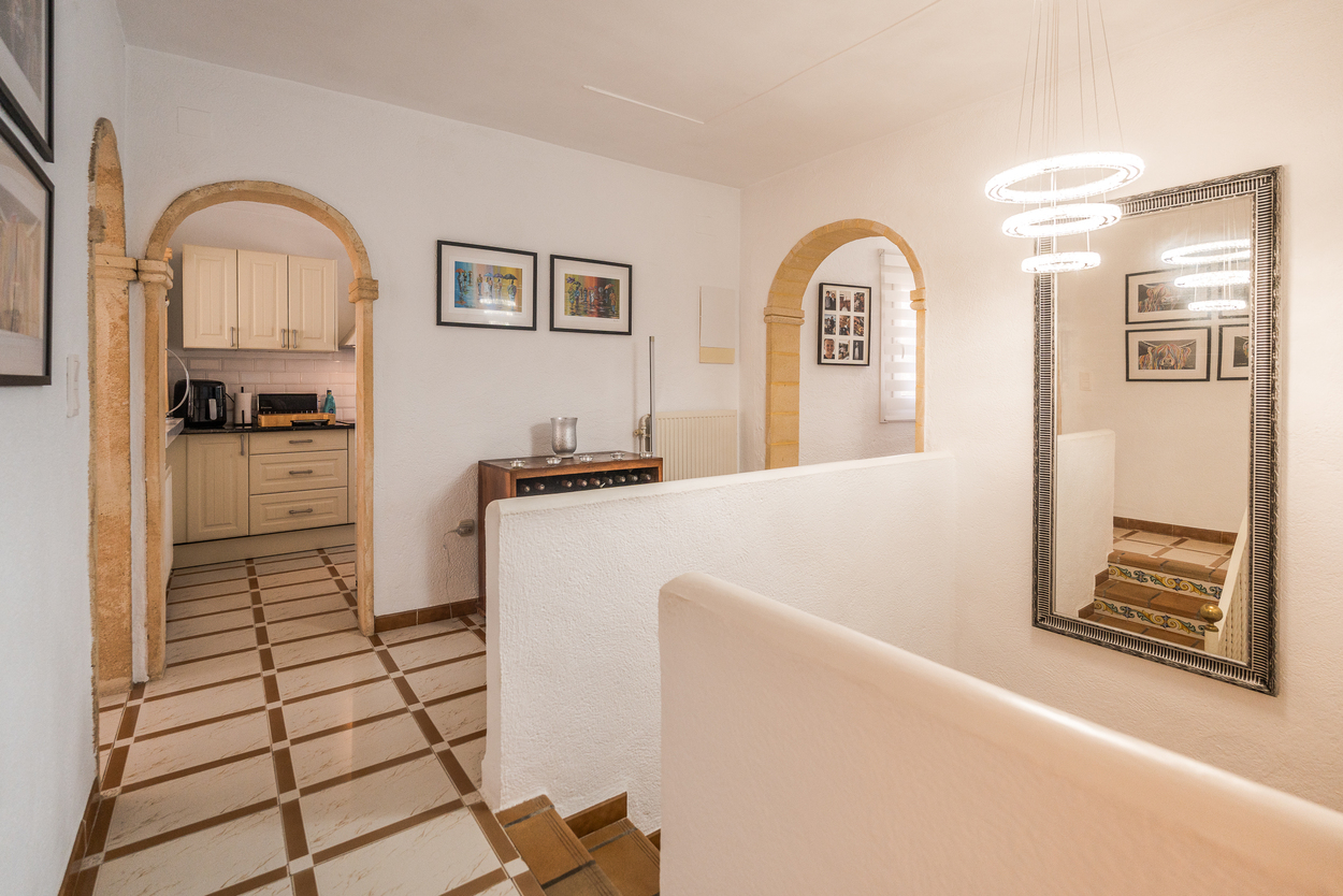 Couloir avec arches en pierre de Tosca, sol carrelé et lustre circulaire moderne. Vue sur la cuisine et l'escalier orné de contremarches décoratives.
