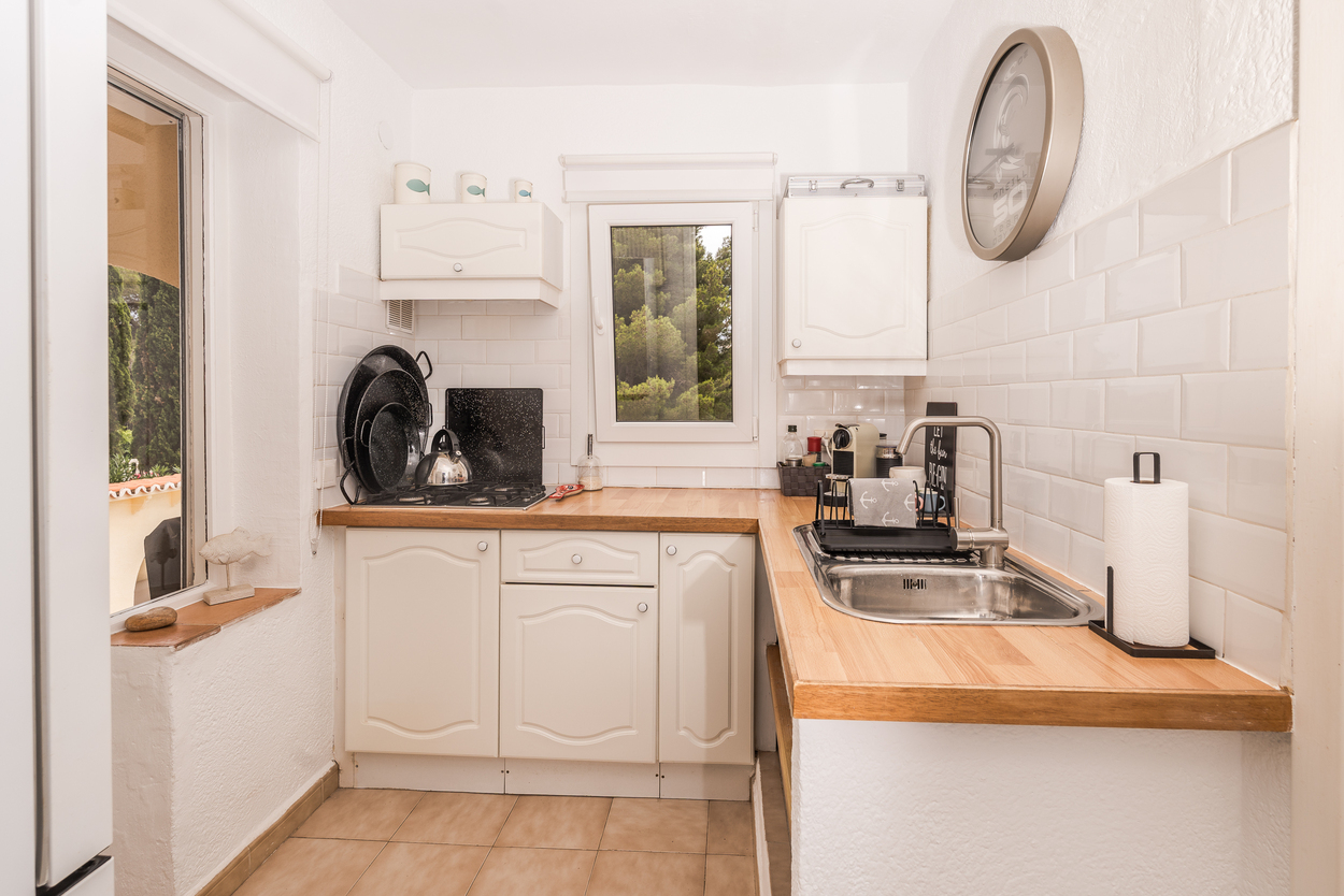 Cuisine compacte avec placards blancs, plans de travail en bois et crédence en carrelage métro. Elle comprend une plaque à gaz, un évier en inox et une vue sur le jardin.