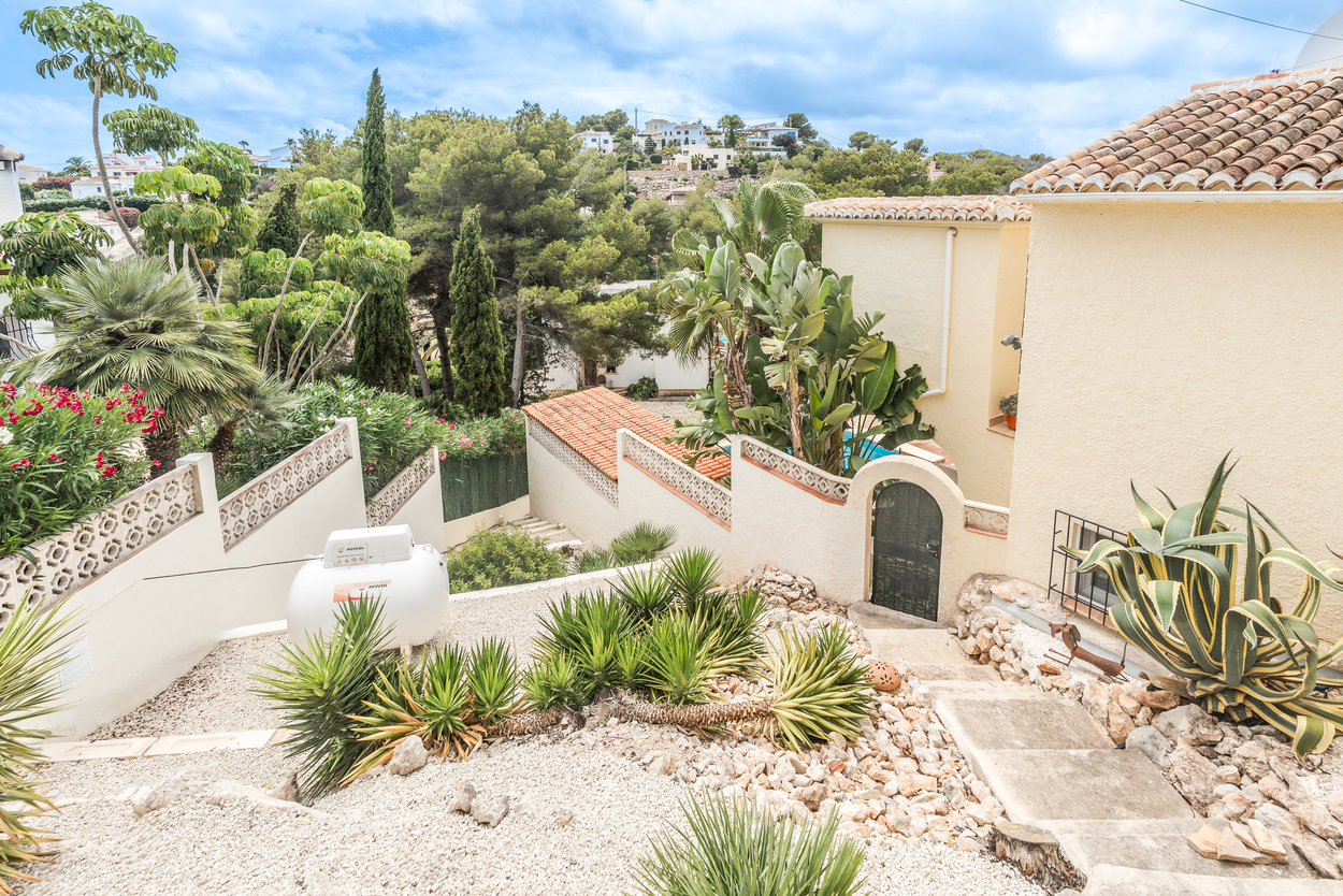 Jardin méditerranéen en terrasses avec gravier blanc, yuccas et agaves. Des marches mènent à une entrée voûtée avec portillon en fer et toitures en tuiles.