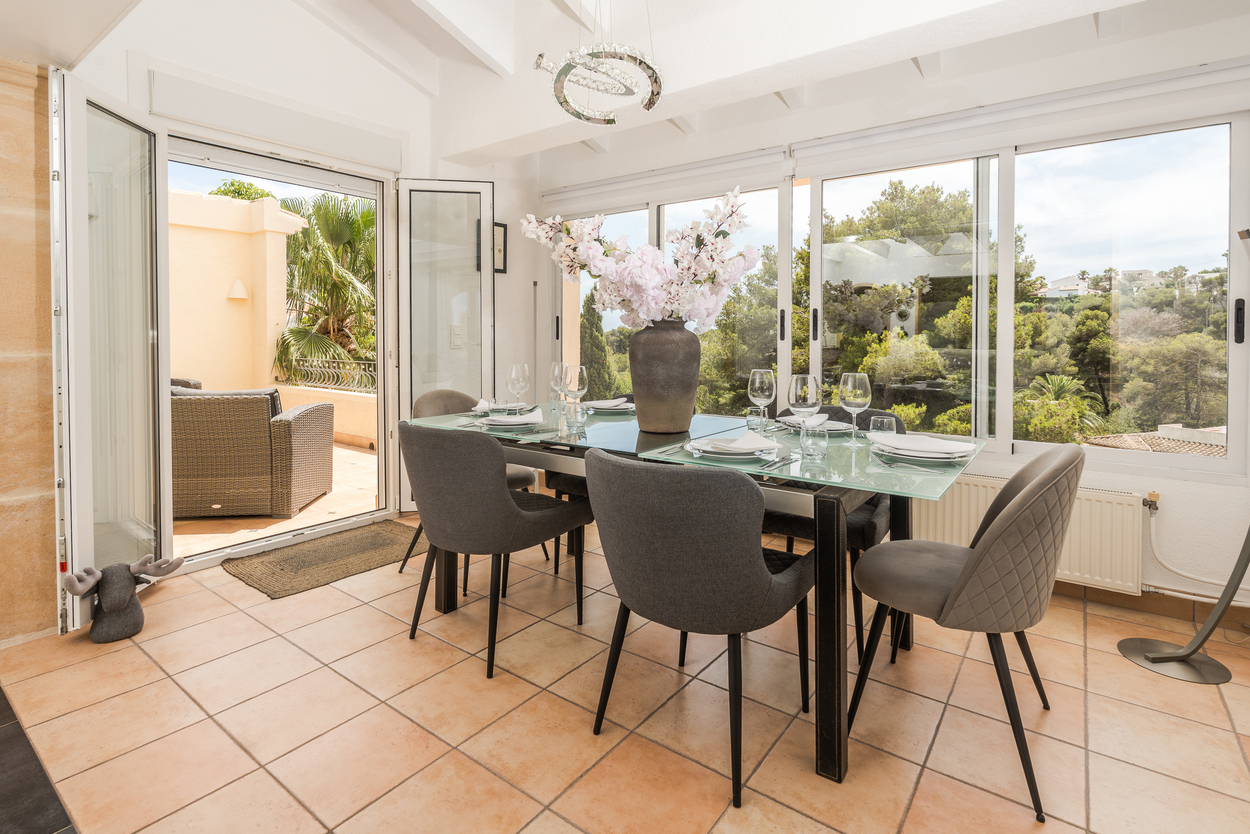 Salle à manger lumineuse avec sol en terre cuite, plafond à poutres blanches et grandes baies vitrées ouvrant sur une terrasse avec vue sur la végétation.