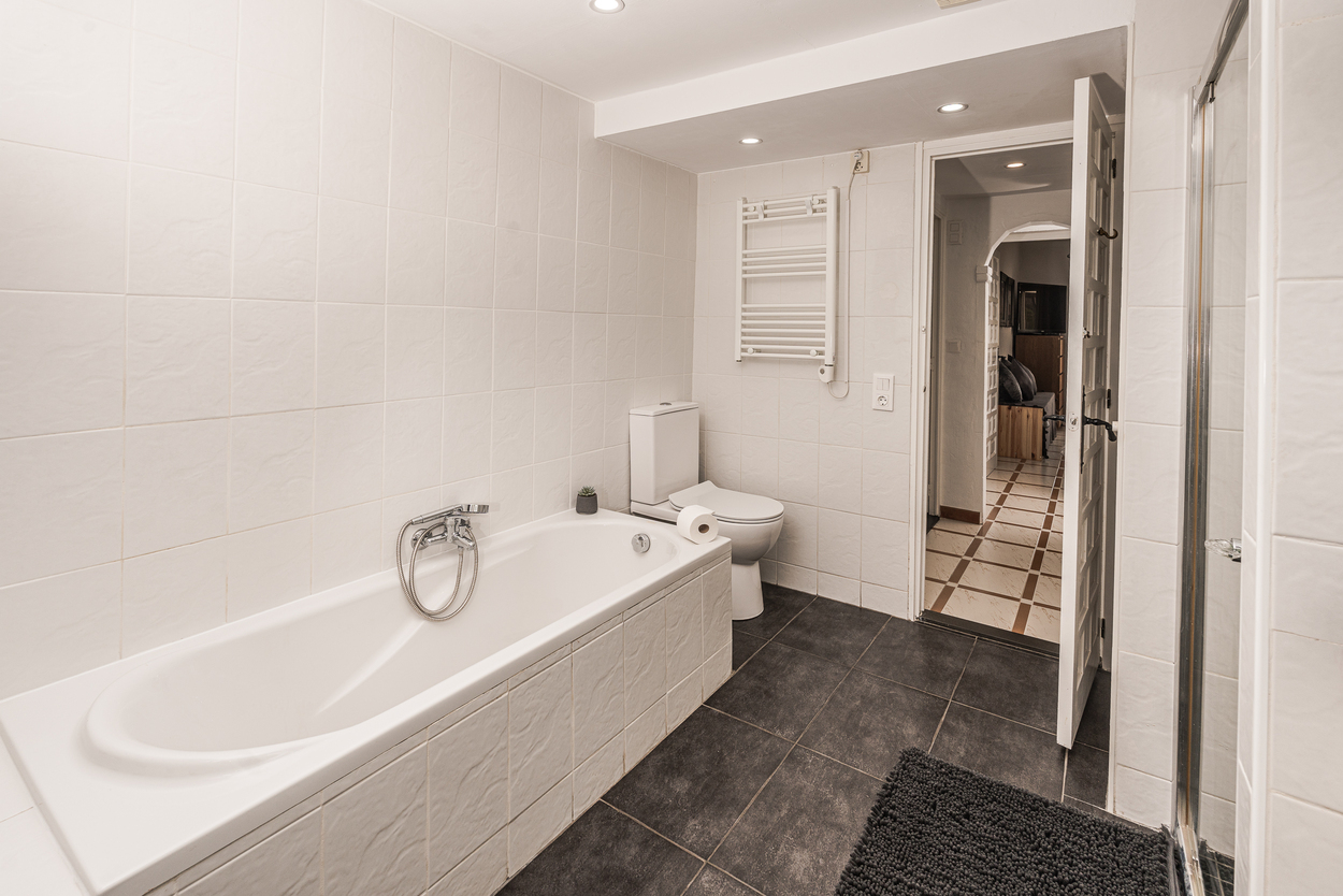 Salle de bain fonctionnelle avec baignoire carrelée en blanc, sol en grès foncé, radiateur sèche-serviettes et spots encastrés.