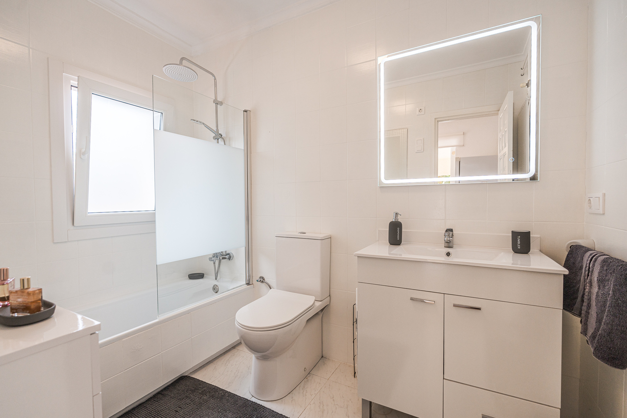 Salle de bain moderne avec carrelage blanc, meuble vasque avec miroir rétroéclairé par LED et baignoire équipée d'une paroi en verre et d'un pommeau de douche.