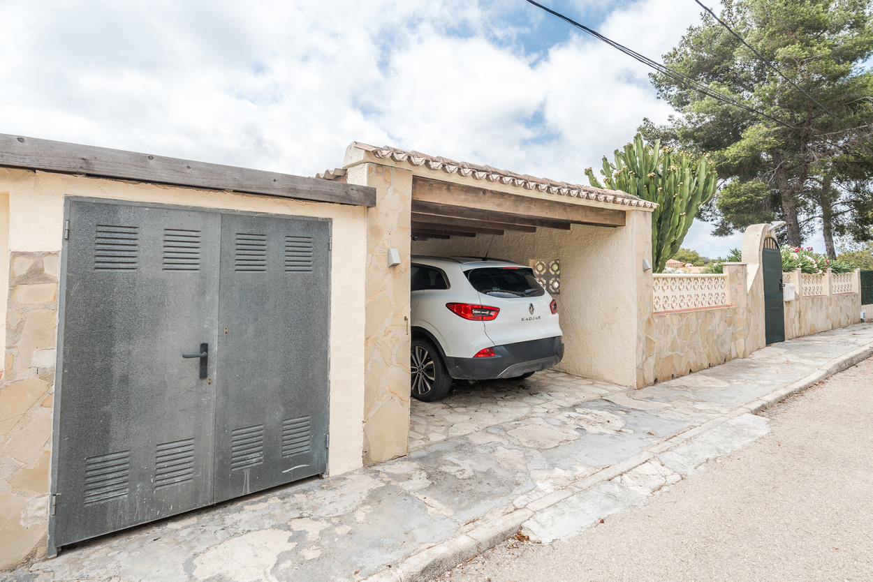 Vue extérieure montrant l'entrée sécurisée, les murs en pierre et un abri de voiture couvert avec poutres en bois et toit en tuiles.