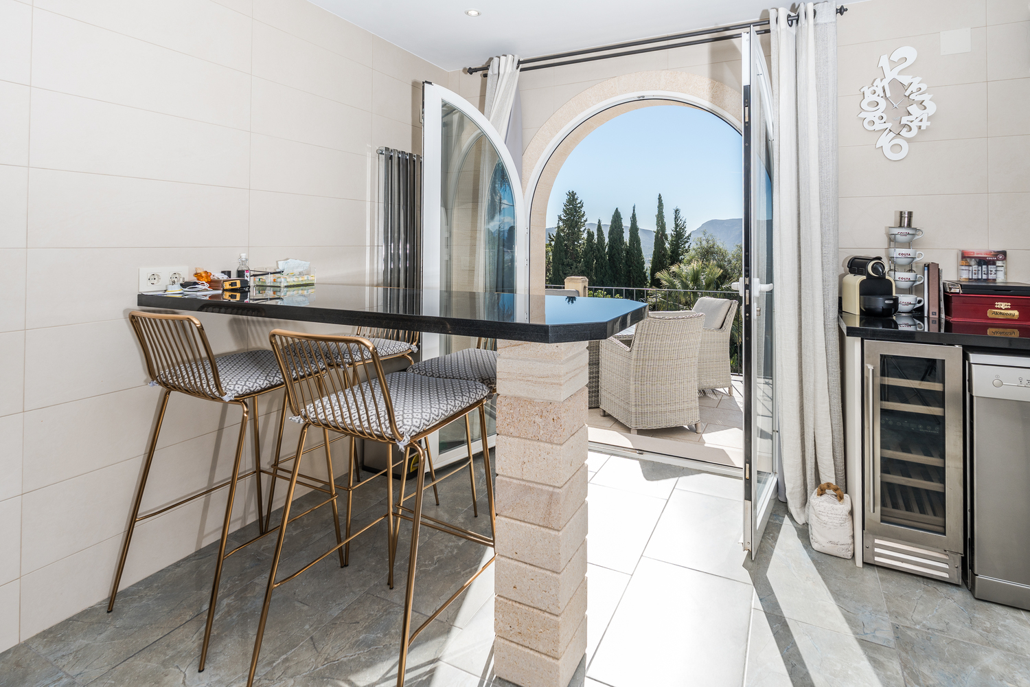 Bar de cuisine avec plan de travail en granit noir et tabourets dorés, ouvrant sur une terrasse via une arche en pierre avec vue sur la vallée et les montagnes.