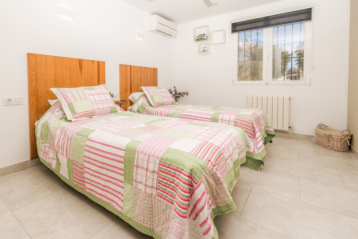 Chambre à deux lits avec têtes de lit en bois, carrelage clair et climatisation. La pièce dispose d'une fenêtre sécurisée et d'un chauffage par radiateur.