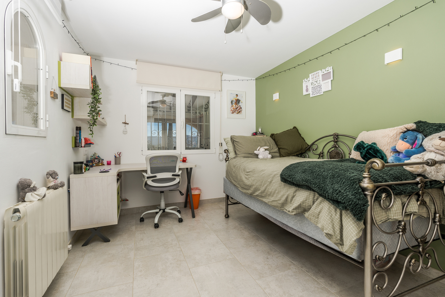 Chambre avec un mur d'accent vert, un lit en métal ornementé et un espace de travail intégré avec bureau et chaise ergonomique. Équipée d'un ventilateur de plafond.