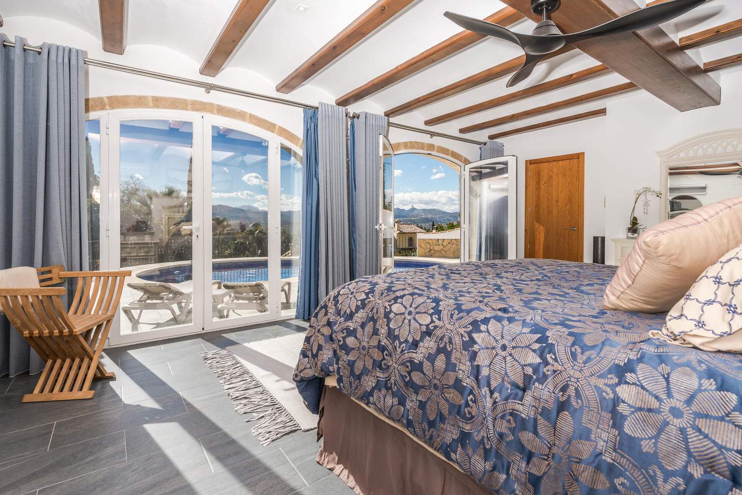 Chambre de maître avec poutres apparentes, arches en pierre et baies vitrées offrant un accès direct à la piscine et une vue dégagée.