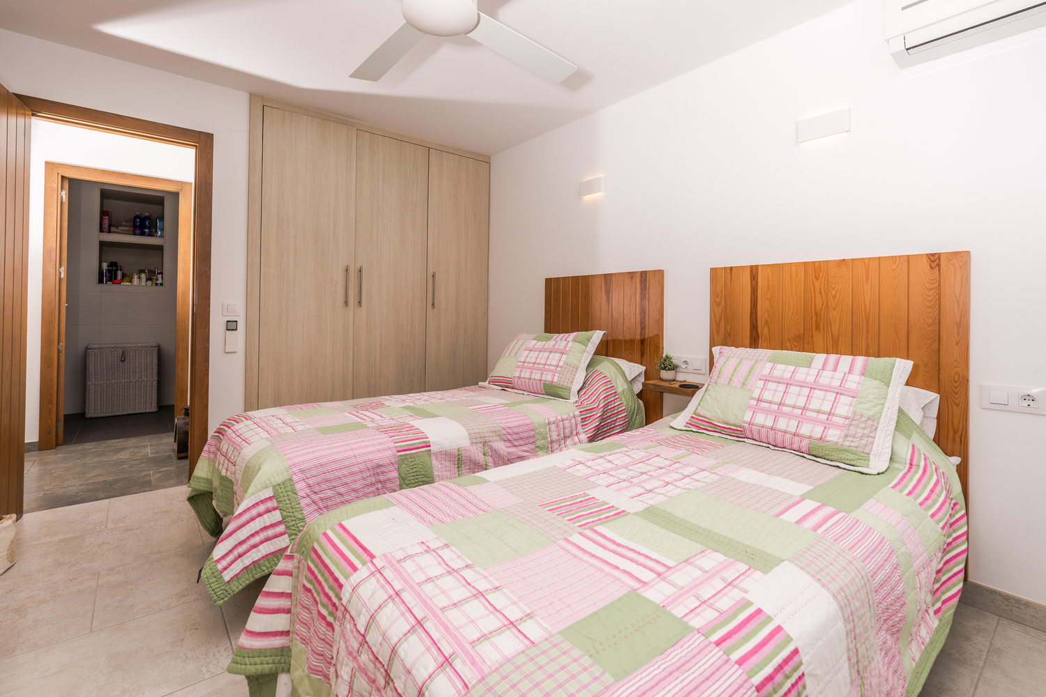 Chambre lumineuse avec lits jumeaux, têtes de lit en bois, placards intégrés, ventilateur de plafond et accès à une salle de bain attenante.