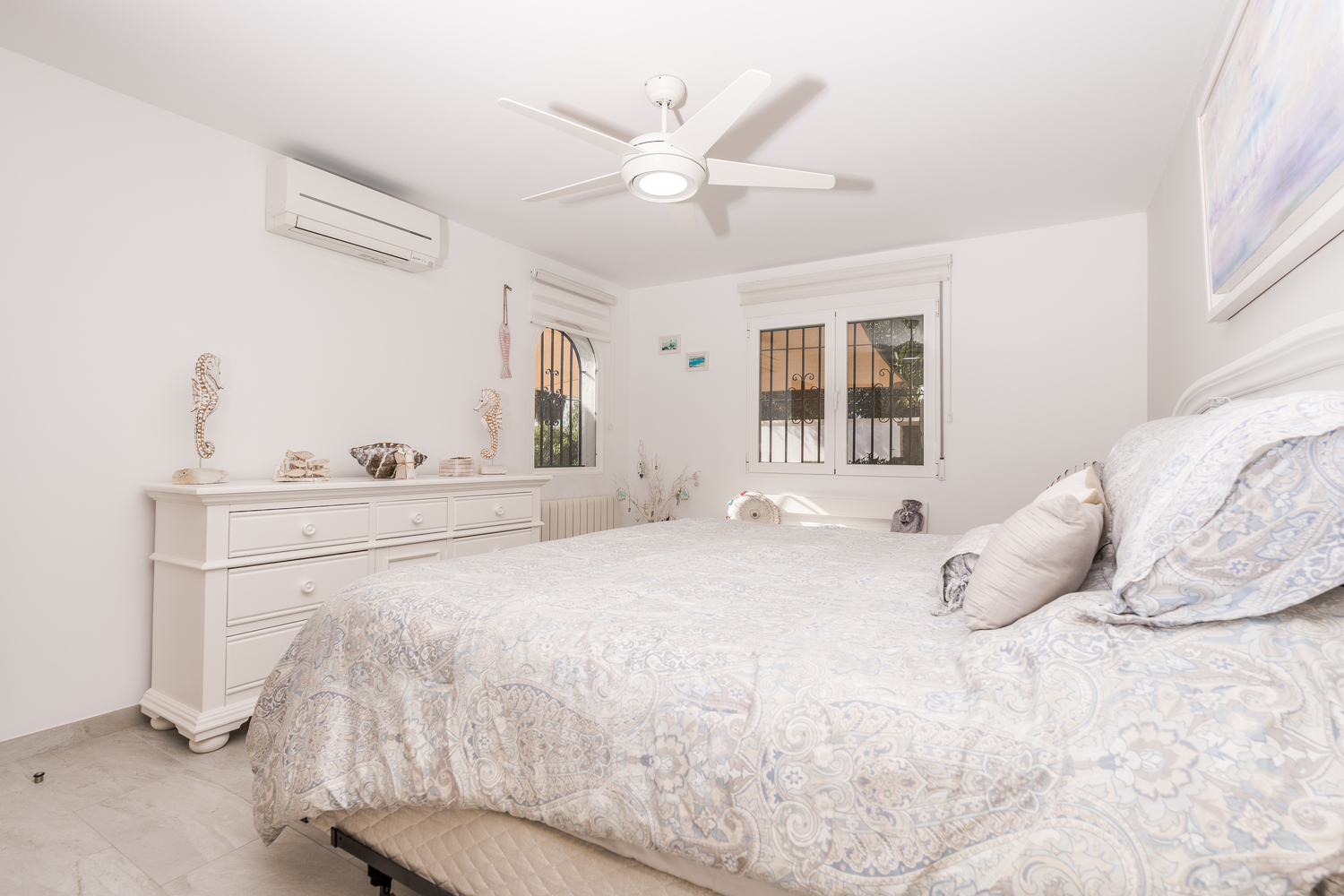 Chambre lumineuse avec murs blancs, ventilateur de plafond, climatisation et commode blanche. Les fenêtres sont équipées de grilles de sécurité et de stores.