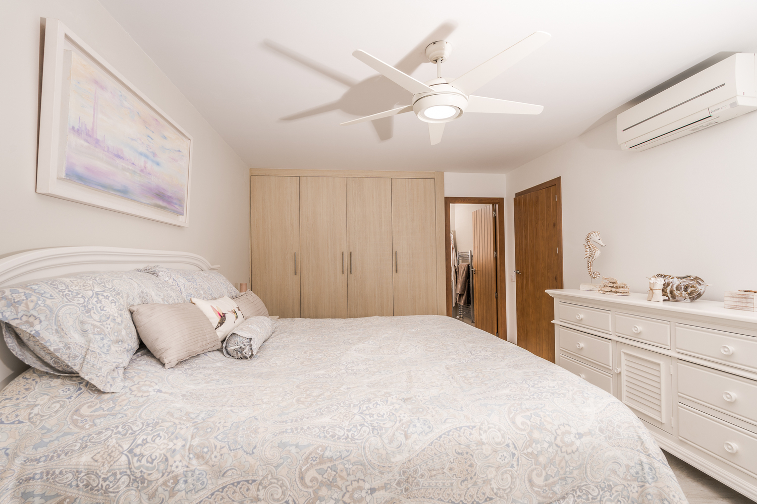 Chambre moderne avec placards intégrés en bois clair, commode blanche et ventilateur de plafond. Équipée d'une climatisation murale et d'un accès à la suite.