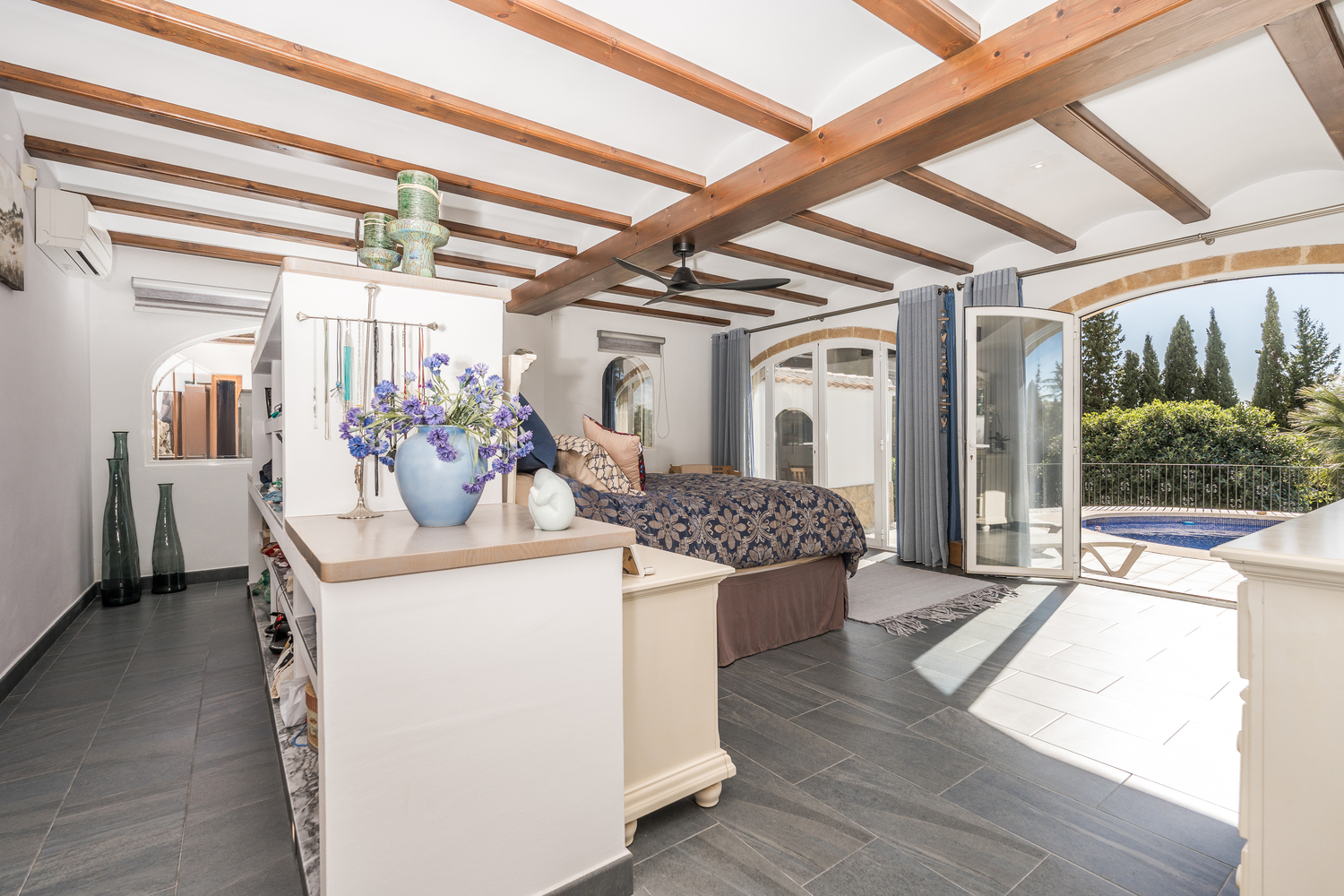 Chambre principale spacieuse avec poutres en bois apparentes, sol en carrelage sombre et portes vitrées voûtées ouvrant directement sur la piscine.
