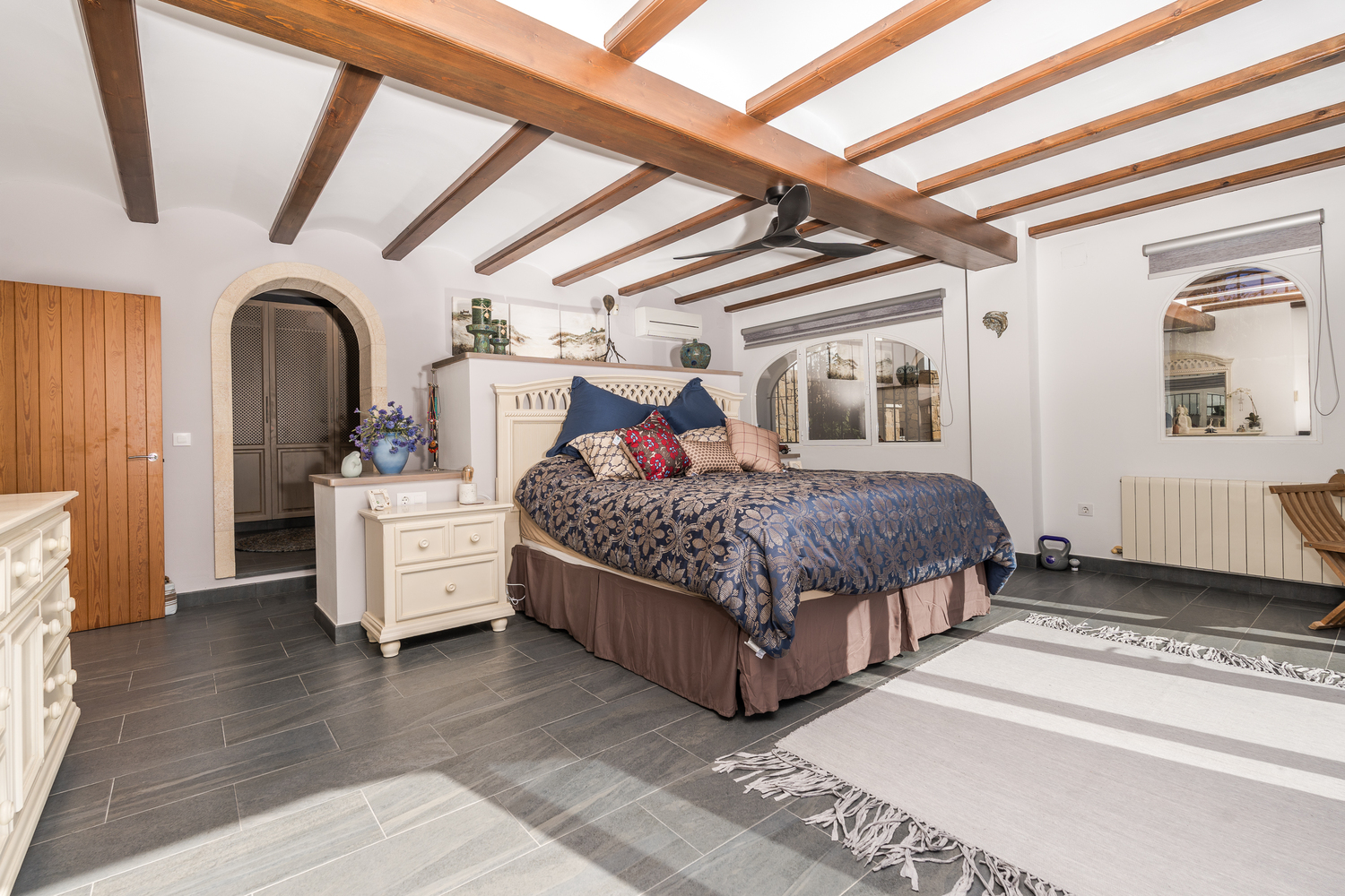 Chambre spacieuse avec poutres en bois apparentes, sol en grès cérame gris et arche en pierre naturelle menant au dressing. Équipée de climatisation et chauffage central.
