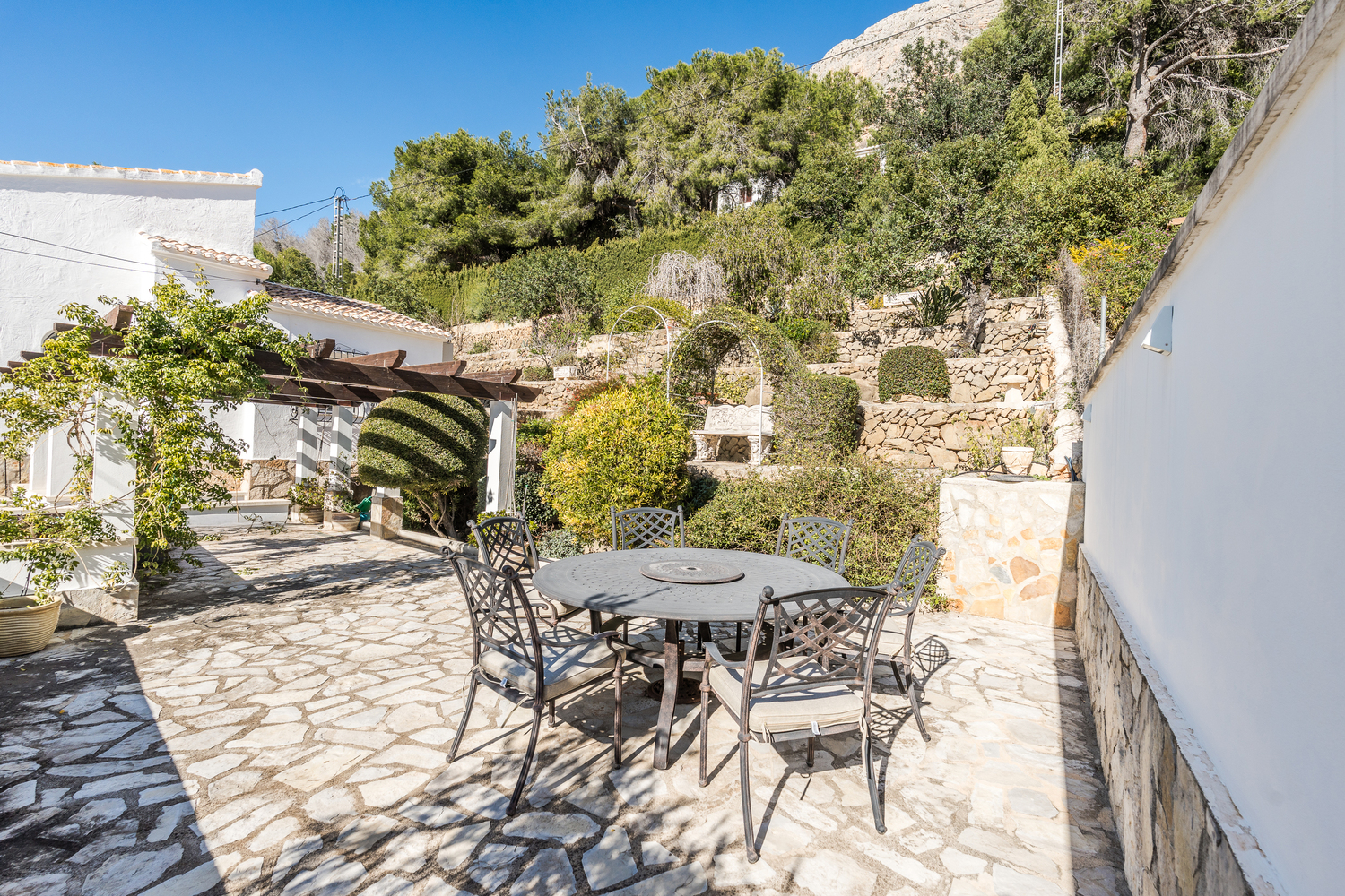 Coin repas extérieur comprenant une table ronde en fer forgé sur un sol en pierre naturelle, avec jardins méditerranéens en terrasses et pergola.