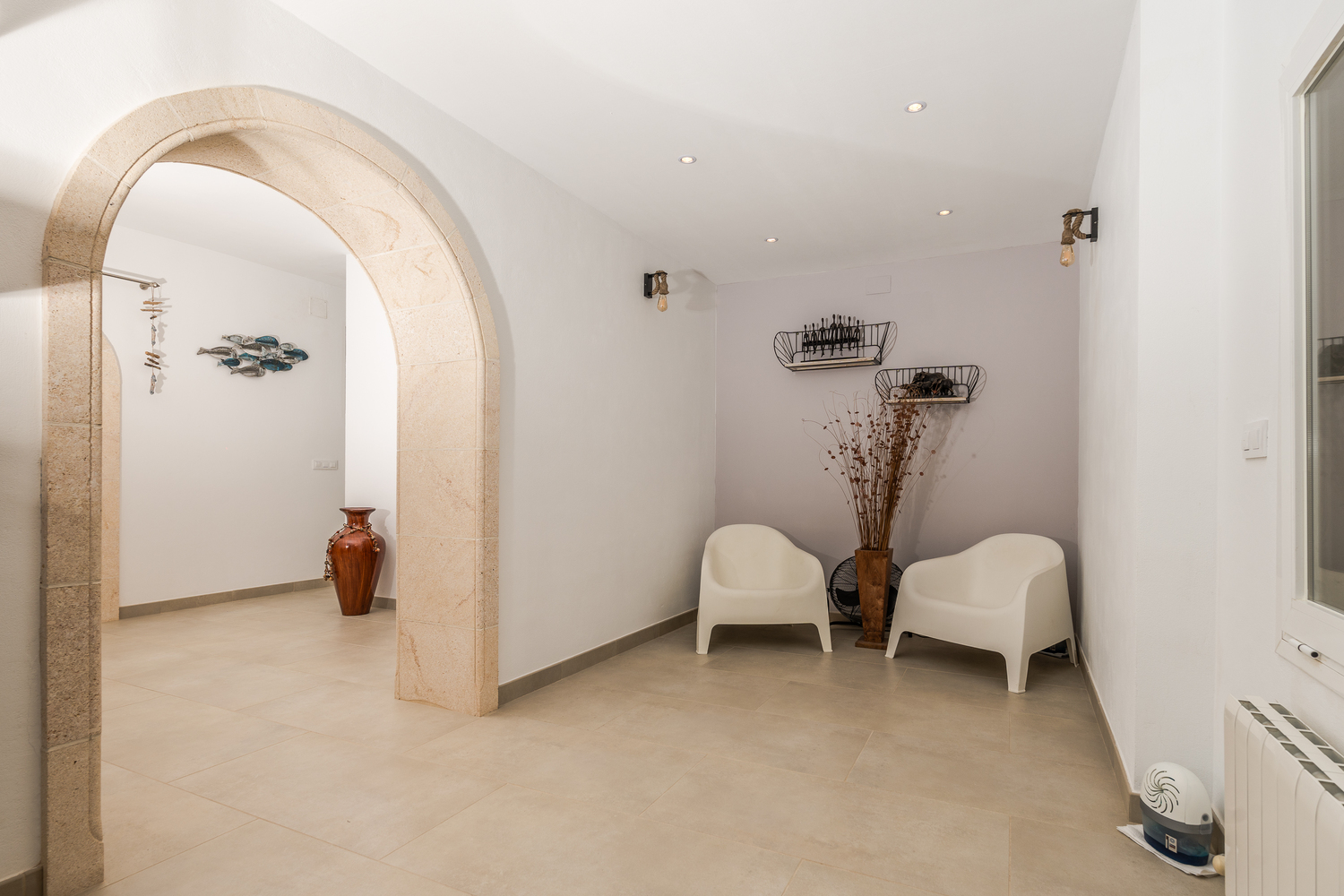 Couloir avec une arche traditionnelle en pierre de Tosca, éclairage encastré et un coin salon minimaliste avec fauteuils blancs et appliques murales industrielles.