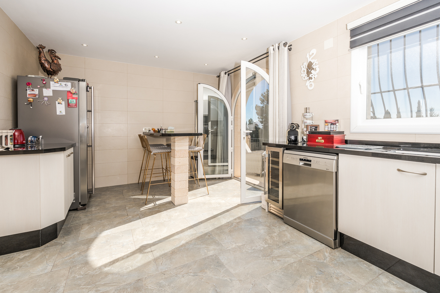 Cuisine moderne avec armoires blanches, plans de travail en granit noir et coin repas. Équipée d'appareils en inox et d'une porte vitrée en arche ouvrant sur la terrasse.
