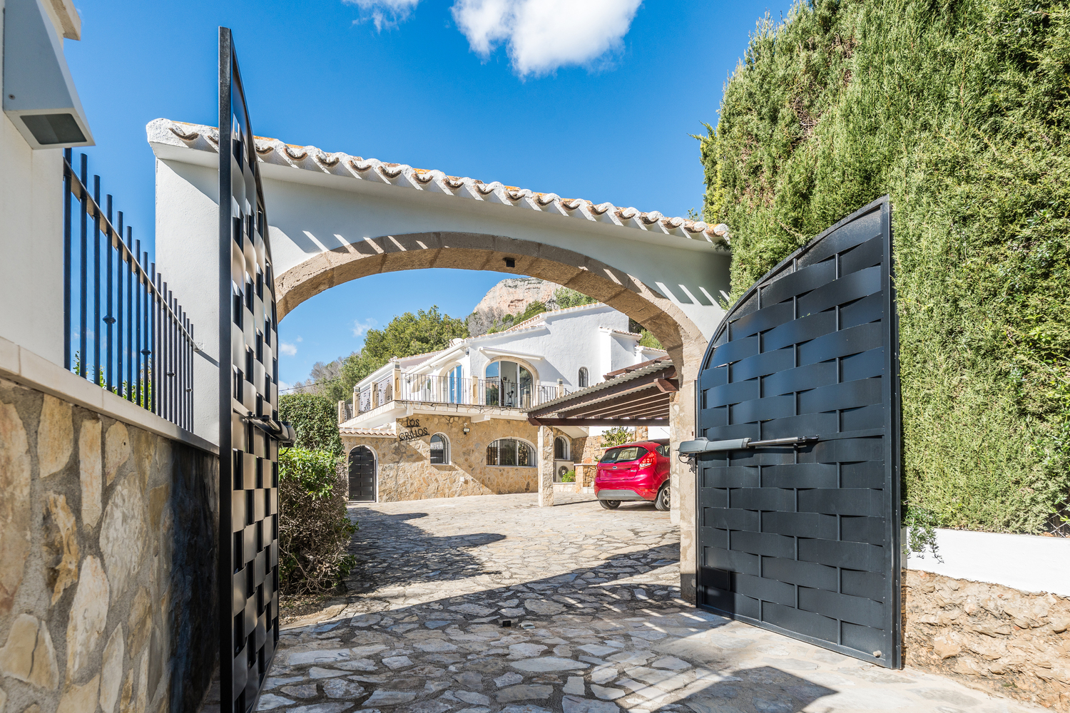 Entrée avec portail automatique en métal noir et arche en pierre de Tosca, s'ouvrant sur une allée pavée menant à la villa et à l'abri de voiture couvert.