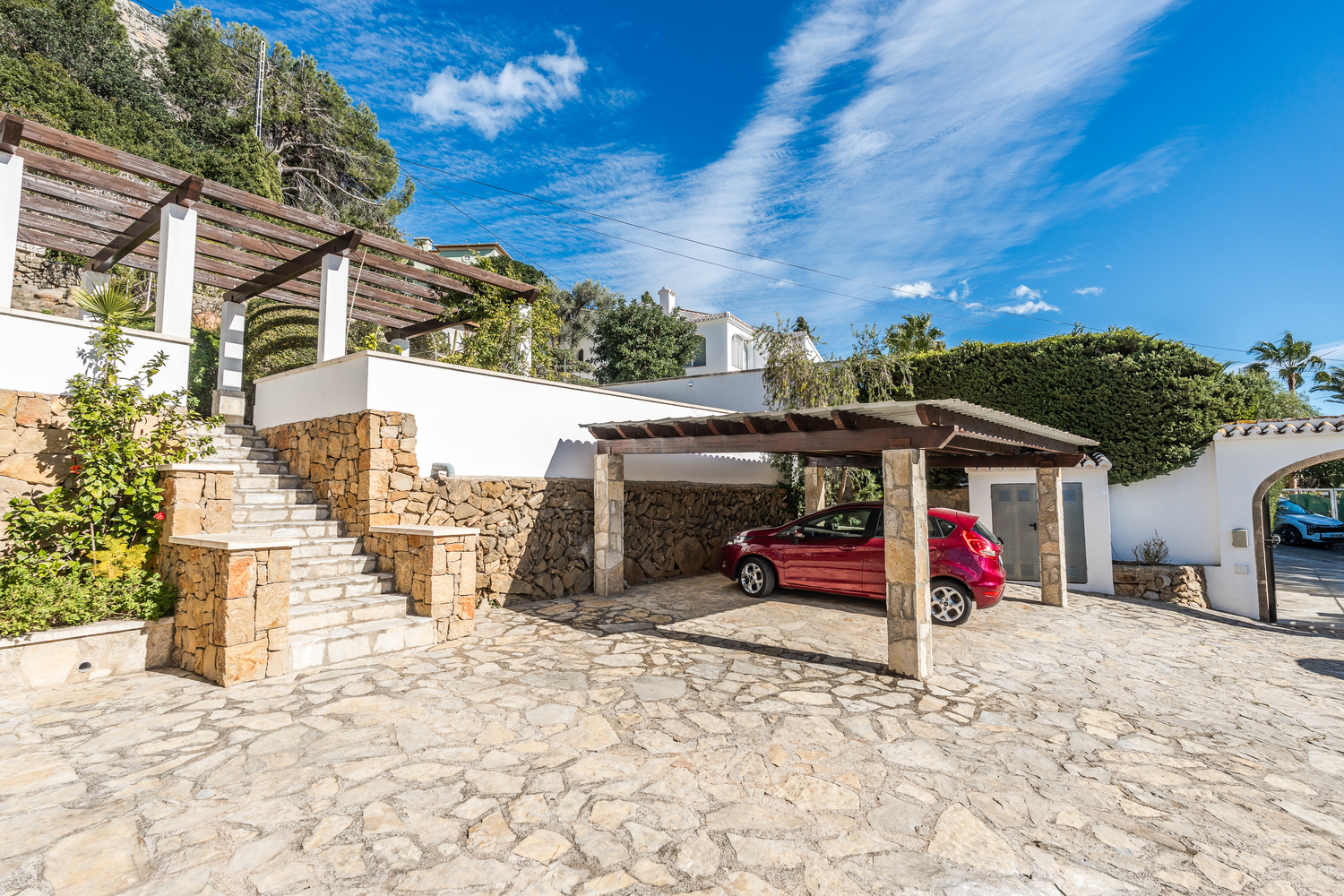 Entrée extérieure avec allée en pierre, abri de voiture couvert avec piliers en pierre et escaliers en maçonnerie menant aux terrasses de la villa.