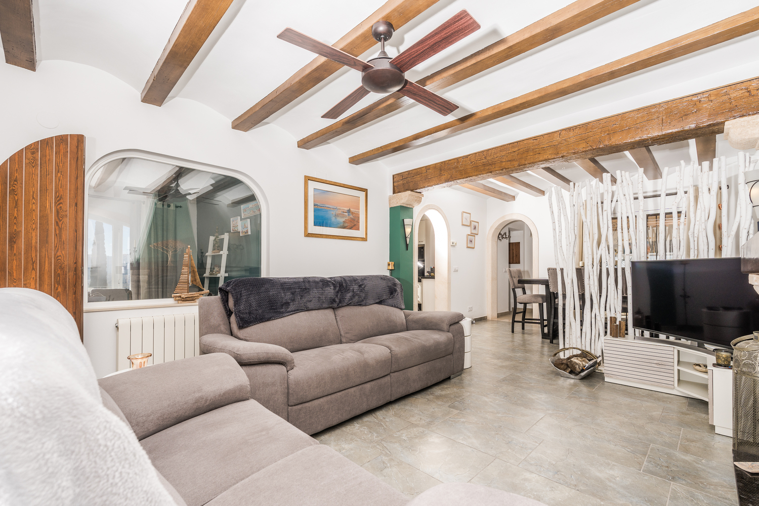 Espace de vie avec poutres en bois apparentes, ventilateur de plafond et carrelage. Une cloison décorative en branches sépare le salon de la salle à manger.