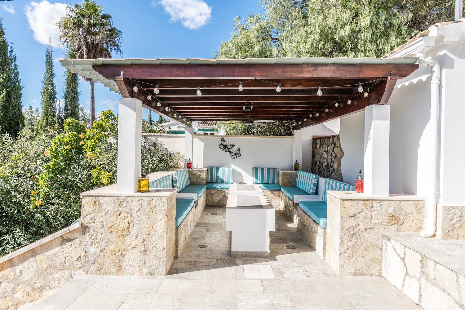 Espace détente extérieur abrité par une pergola en bois avec ventilateur, banquettes en pierre intégrées et coussins rayés bleu turquoise.