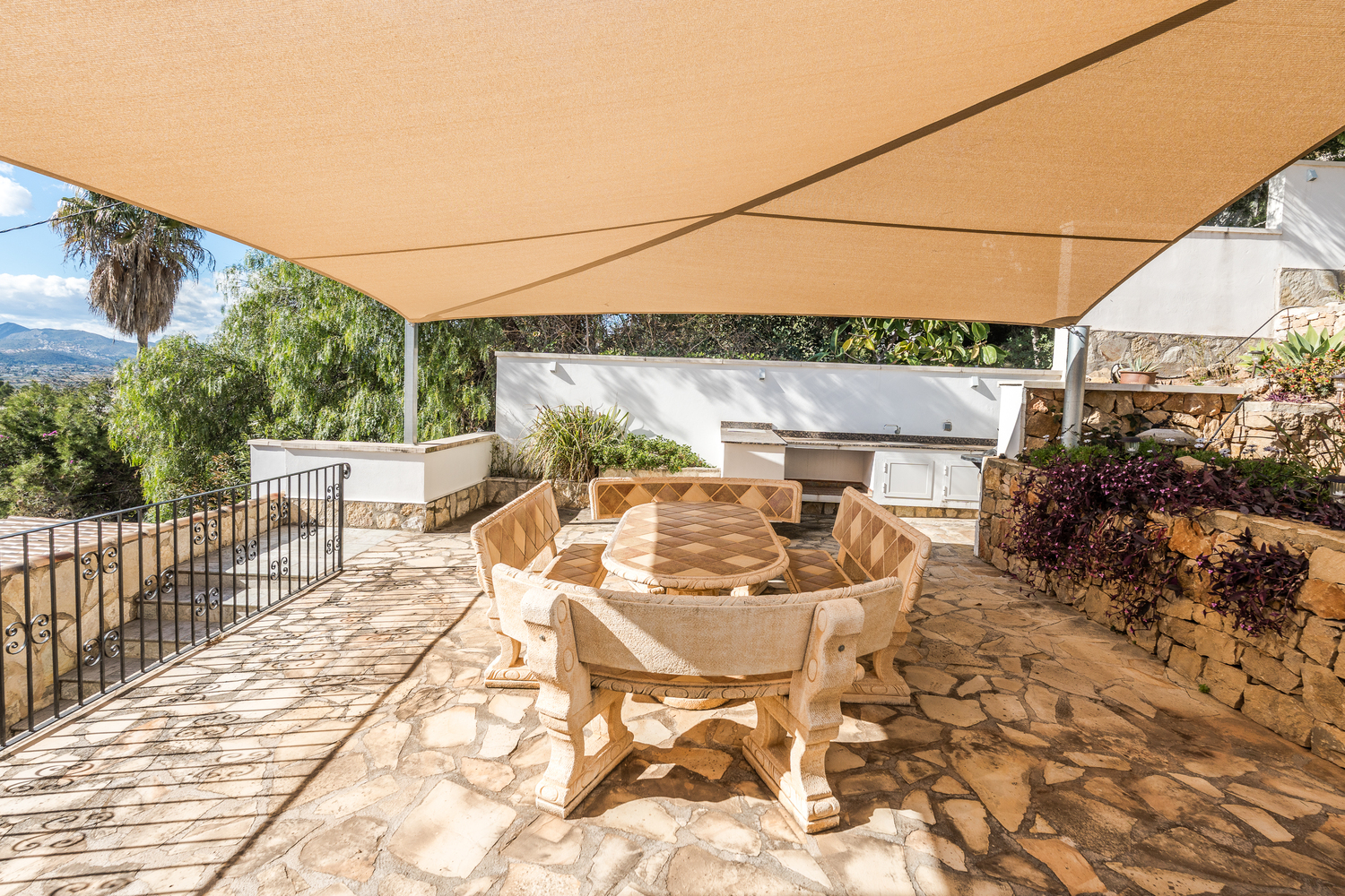 Espace repas extérieur avec table et bancs en pierre sculptée, ombragé par une voile d'ombrage beige sur une terrasse en dalles de pierre naturelle.
