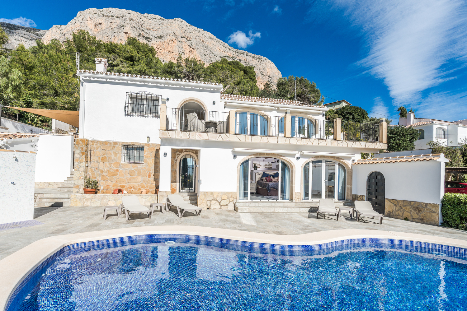Extérieur de la villa avec piscine en mosaïque, terrasse en pierre avec transats et architecture méditerranéenne sur fond de montagne Montgó.