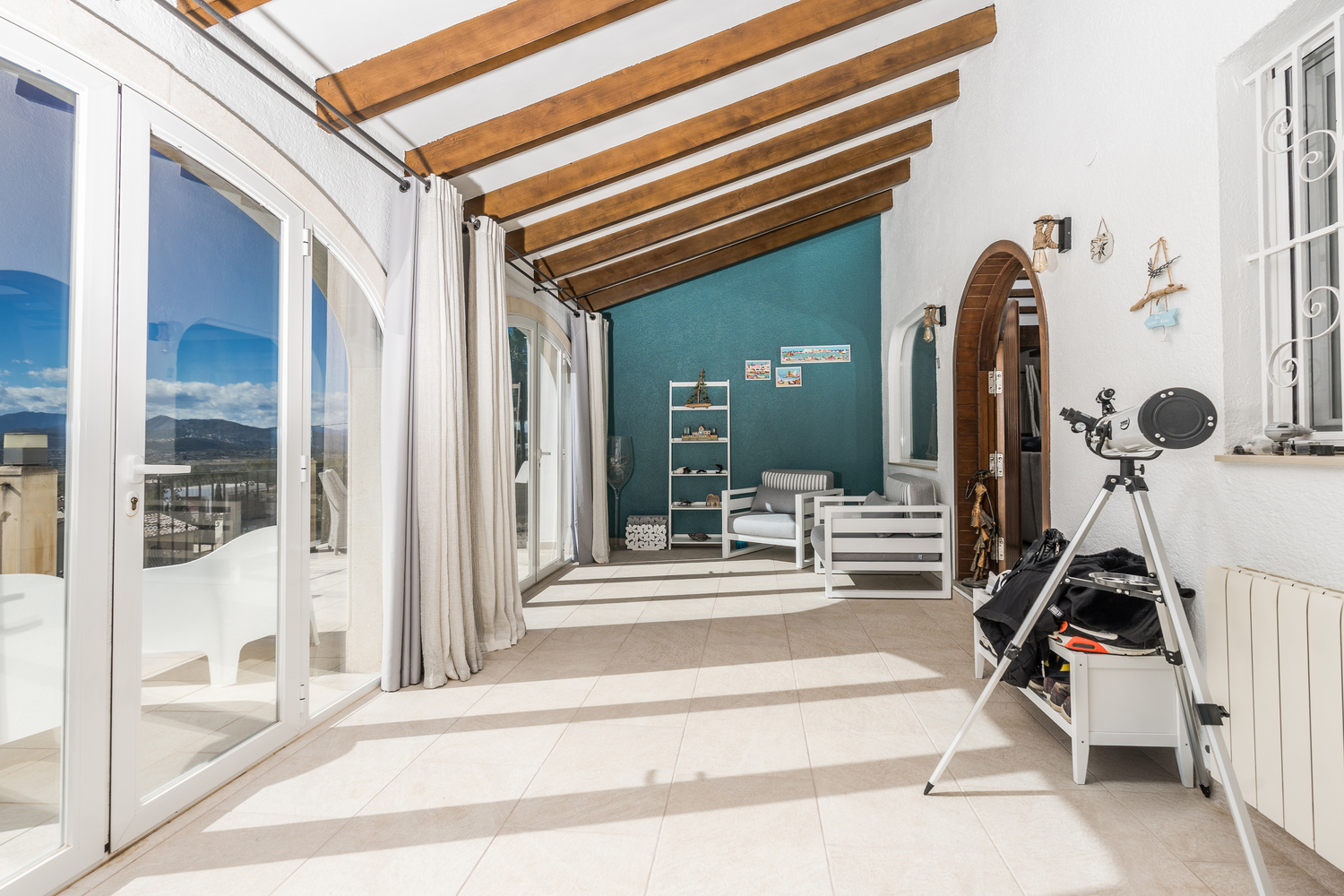 Naya vitrée avec poutres en bois apparentes, sol en carrelage et grandes baies vitrées offrant une vue panoramique sur les montagnes environnantes.