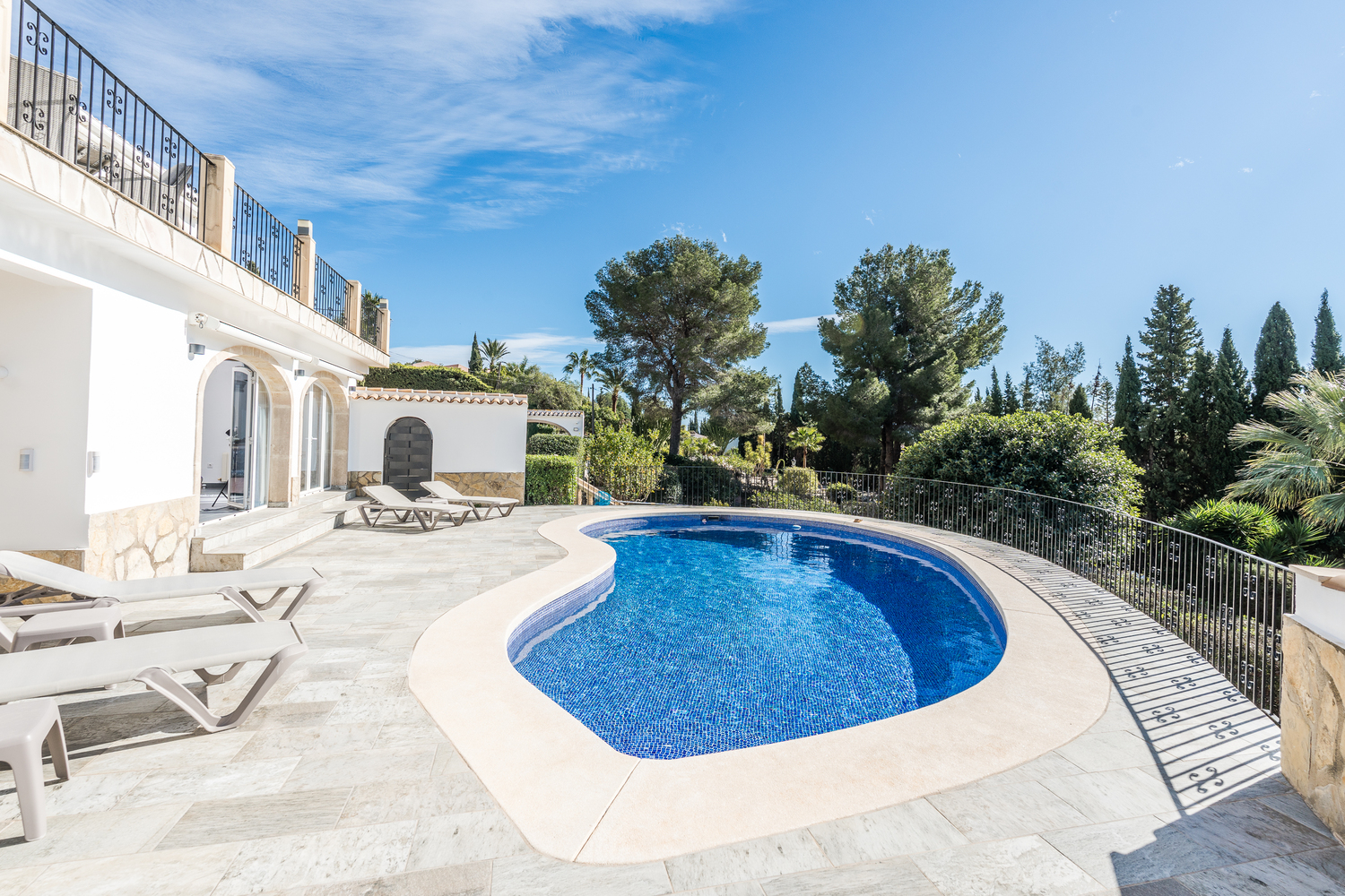 Piscine en forme de rein entourée d'une terrasse en pierre claire avec chaises longues, bordée d'arbres méditerranéens et de la façade blanche de la villa.