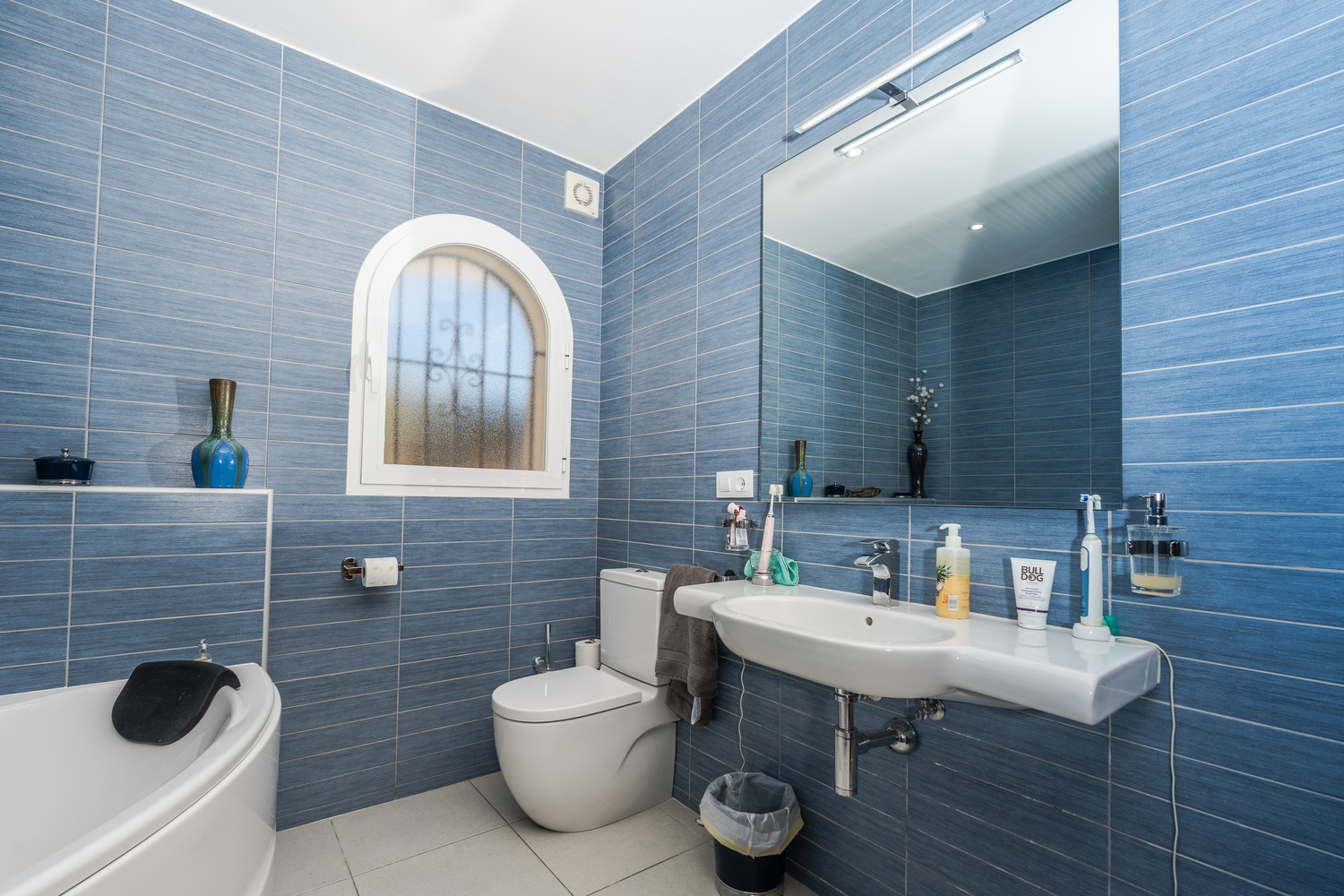 Salle de bain moderne avec carrelage bleu texturé, fenêtre en arc avec verre dépoli, lavabo blanc et un grand miroir avec éclairage intégré.