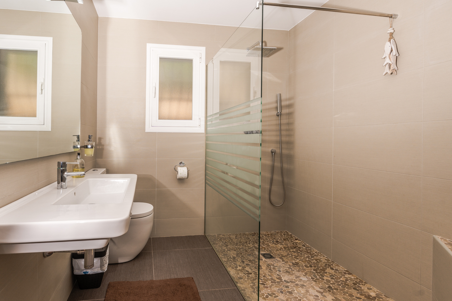 Salle de bain moderne avec douche à l'italienne, sol en galets, paroi en verre et carrelage aux tons neutres. Comprend un lavabo suspendu et une fenêtre dépolie.