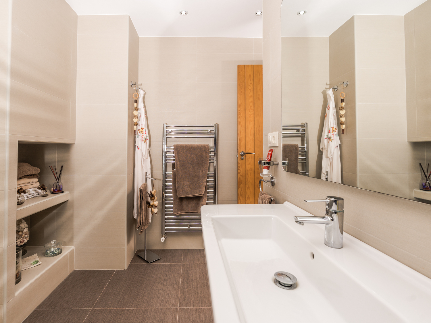 Salle de bain moderne avec une grande vasque en céramique, des étagères murales encastrées et un sèche-serviettes chromé sur carrelage beige.