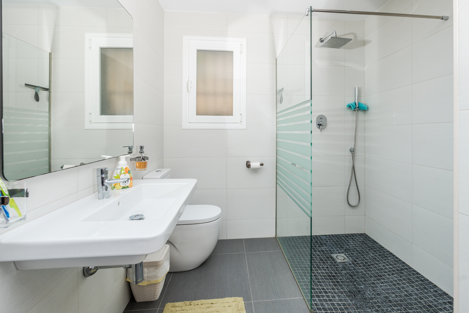 Salle de bains moderne avec douche à l'italienne, paroi vitrée et sol en mosaïque foncée. Elle dispose d'un lavabo suspendu et d'un carrelage blanc lumineux.