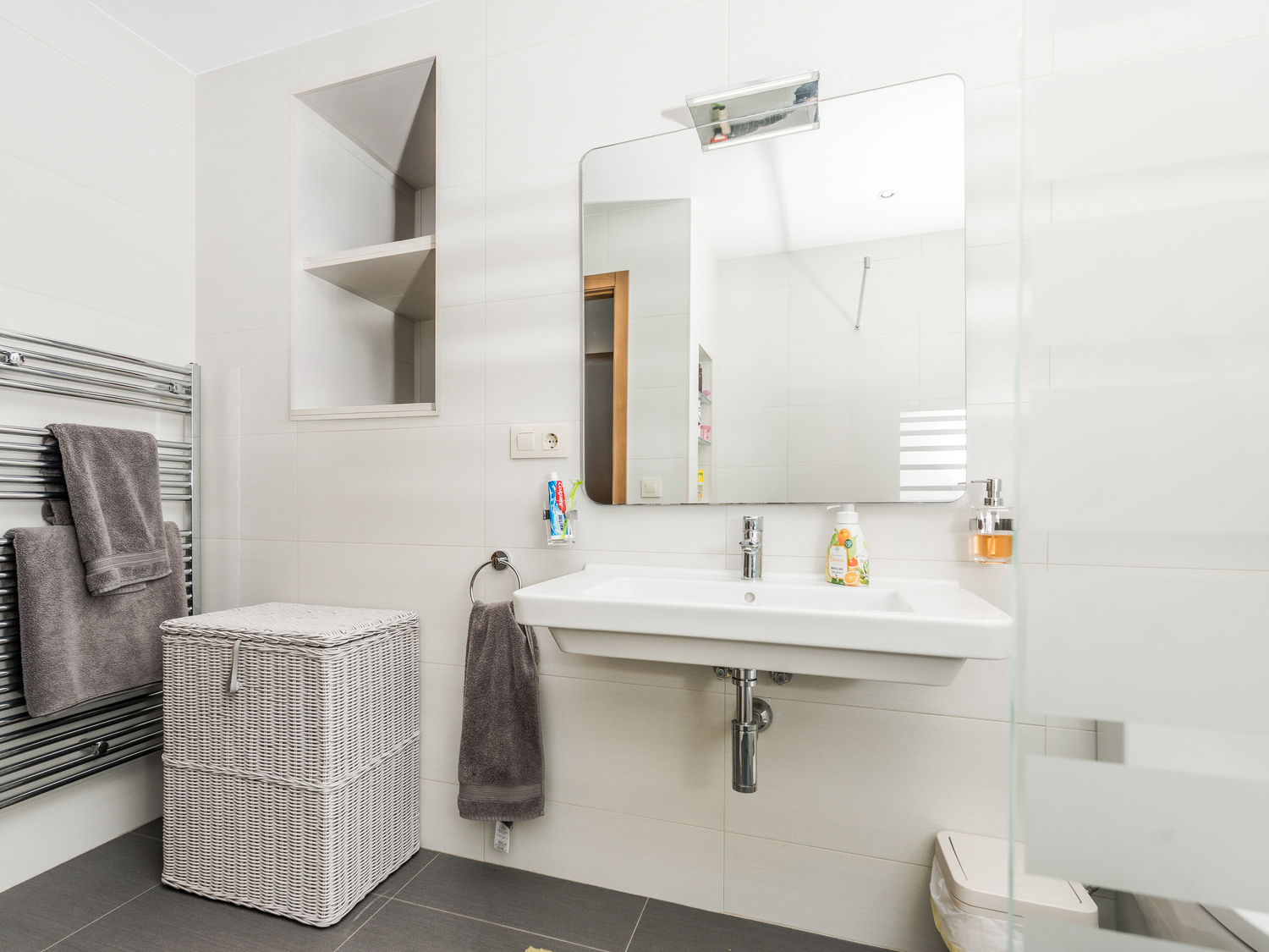 Salle de bains moderne avec lavabo suspendu, niche murale intégrée et sèche-serviettes chromé sur carrelage blanc et sol gris anthracite.