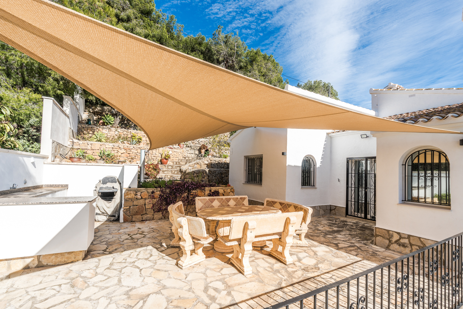Terrasse de salle à manger extérieure avec mobilier en pierre sculptée sous une voile d'ombrage, entourée de murs en pierre sèche et de plantes méditerranéennes.