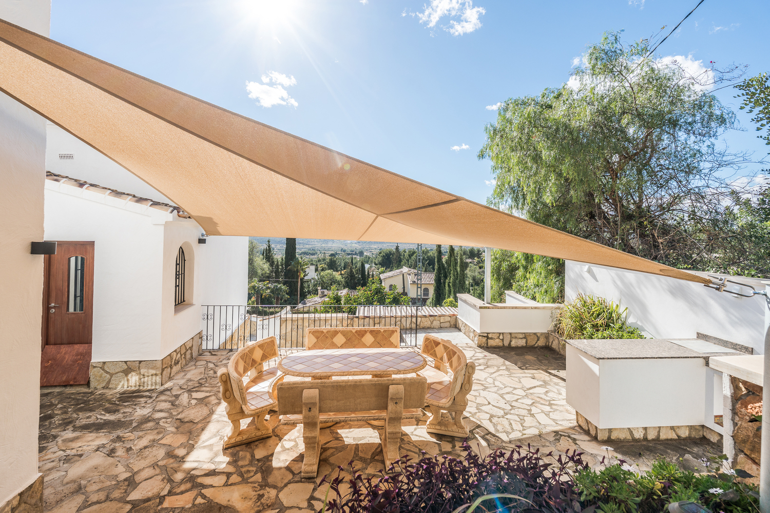 Terrasse extérieure dotée d'un mobilier en pierre sous une voile d'ombrage, offrant une vue panoramique sur la vallée et un coin cuisine d'été.