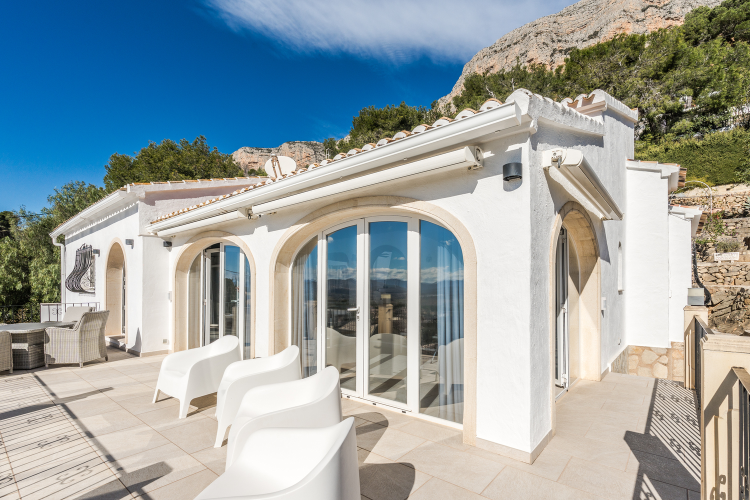 Terrasse supérieure avec naya vitrée, arches en pierre naturelle, mobilier moderne et stores bannes au pied de la montagne Montgó.