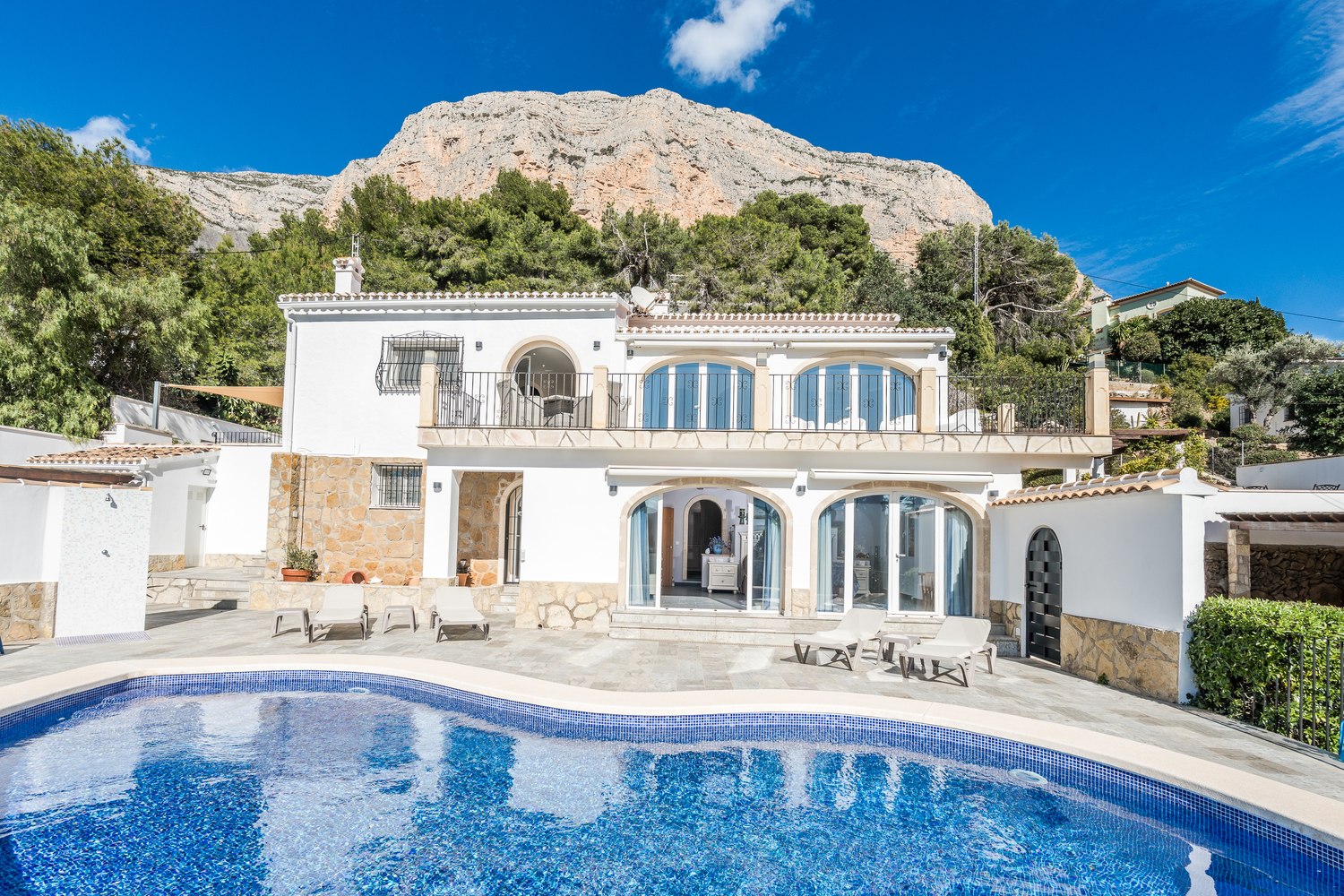 Vue de la façade avec piscine de forme libre, terrasse pavée et ouvertures cintrées, située au pied des falaises calcaires du Montgó.