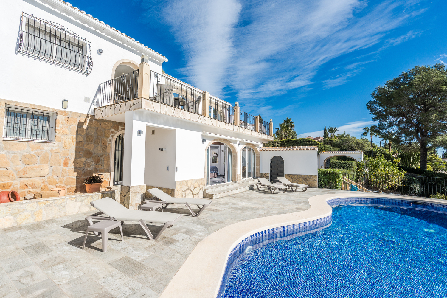 Vue extérieure de la piscine en mosaïque incurvée et de la terrasse en pierre, avec chaises longues et façade blanche ornée de pierre naturelle et d'arches.