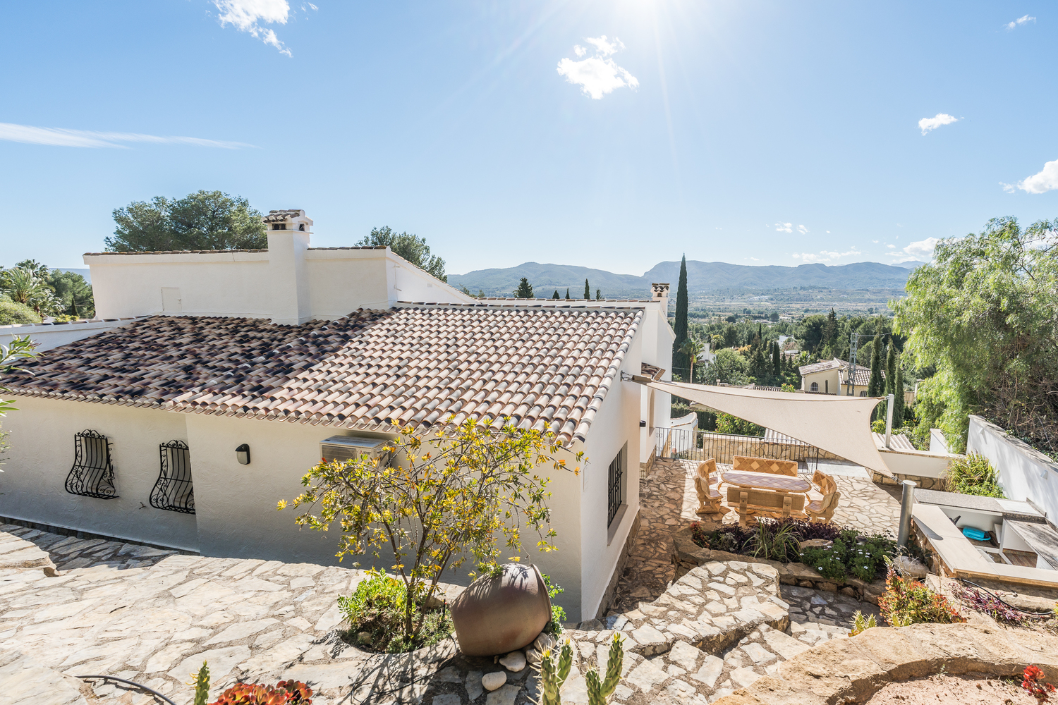 Vue extérieure de la villa avec toiture en terre cuite, terrasses en pierre, coin repas ombragé par une voile d'ombrage et vue panoramique sur les montagnes.