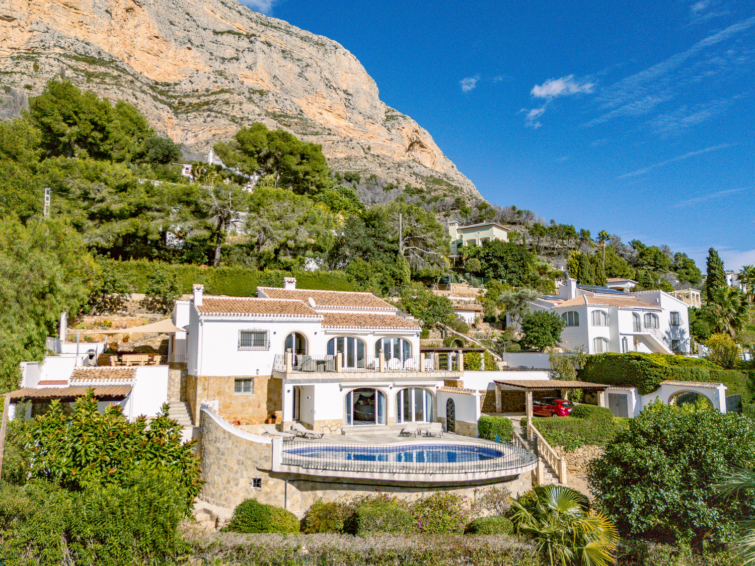Vue extérieure de la villa sur deux niveaux au pied du Montgó, comprenant une piscine courbe, des façades blanches avec soubassements en pierre et des terrasses voûtées.