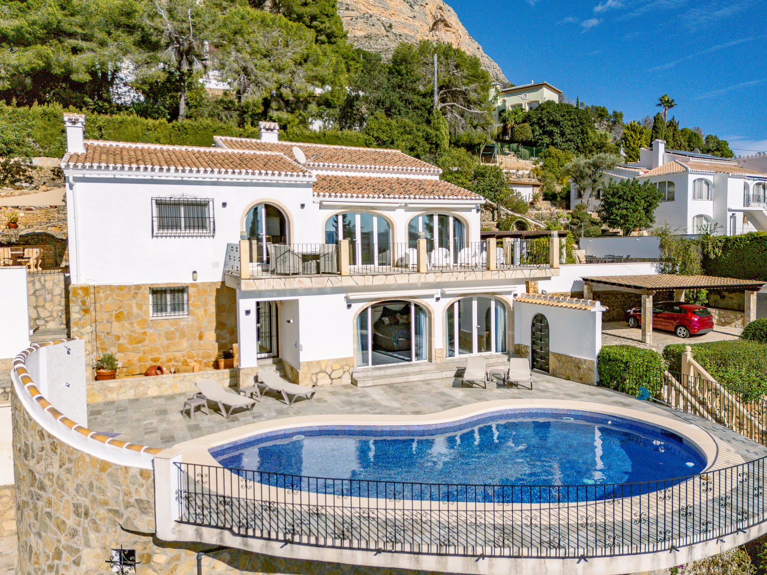 Vue extérieure de la villa sur deux niveaux avec piscine en forme de rein, terrasse en pierre et menuiseries en arche au pied du Montgó.