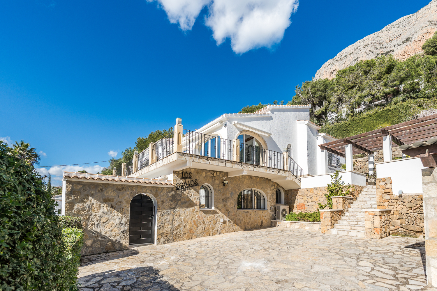 Vue extérieure présentant la façade en pierre naturelle, les fenêtres cintrées et une grande allée pavée au pied de la montagne Montgó.