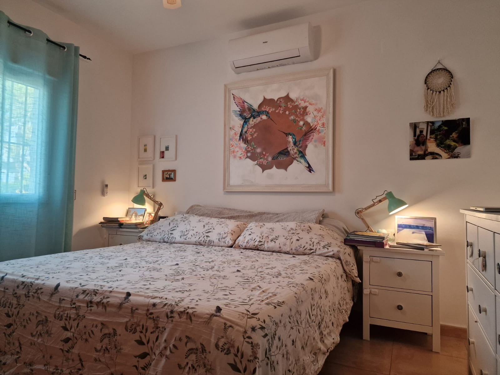 Chambre lumineuse avec lit double, climatisation Haier, tables de chevet avec lampes vertes et une grande œuvre d'art représentant des colibris au-dessus du lit.