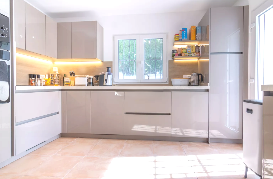 Cuisine moderne avec des meubles taupe brillant, des plans de travail blancs et un éclairage LED intégré. Sol en carrelage de terre cuite et appareils encastrés.