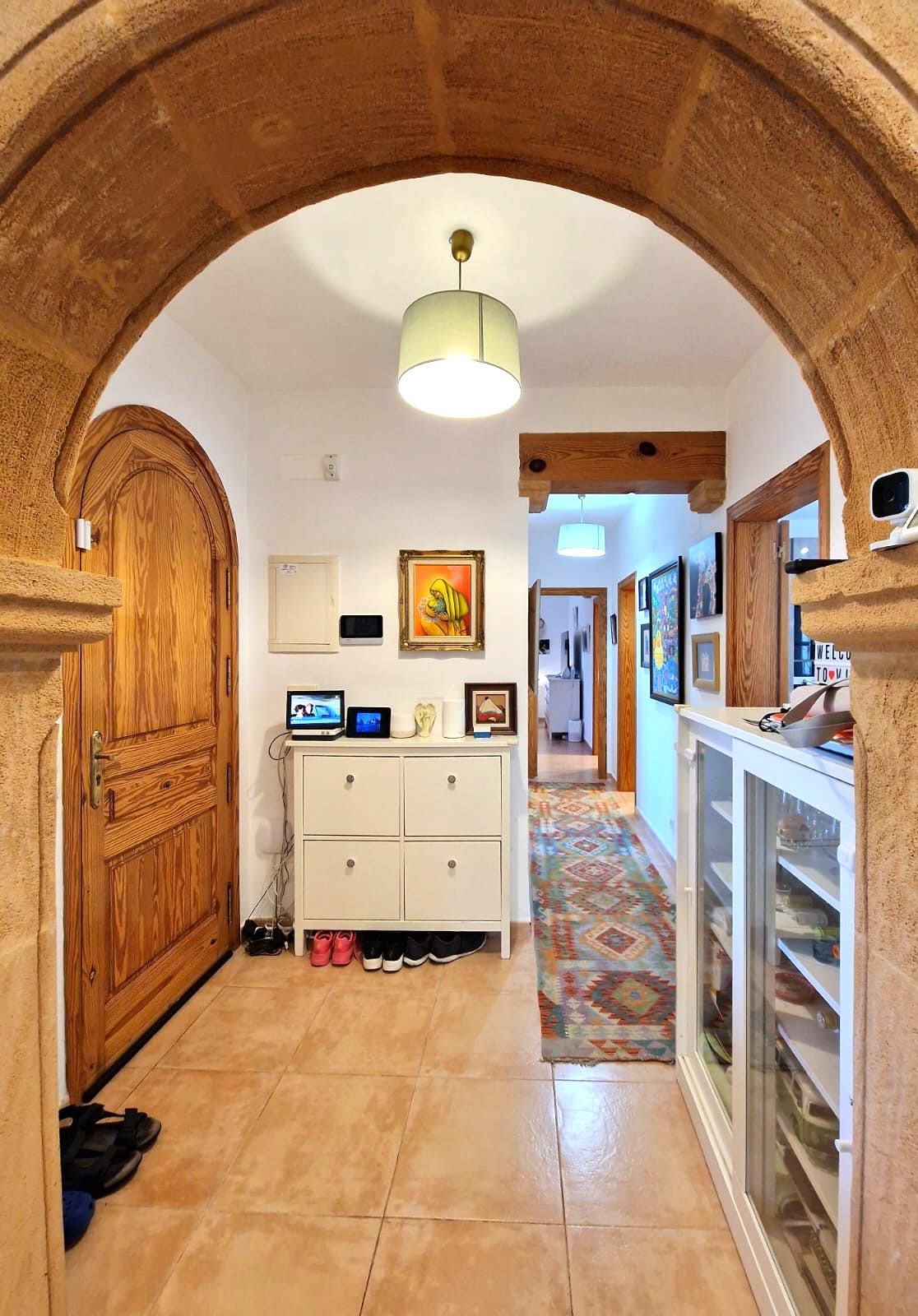 Entrée vue à travers une arche en pierre Tosca, comprenant une porte en bois voûtée, un sol carrelé et un couloir avec un tapis à motifs et des poutres en bois apparentes.
